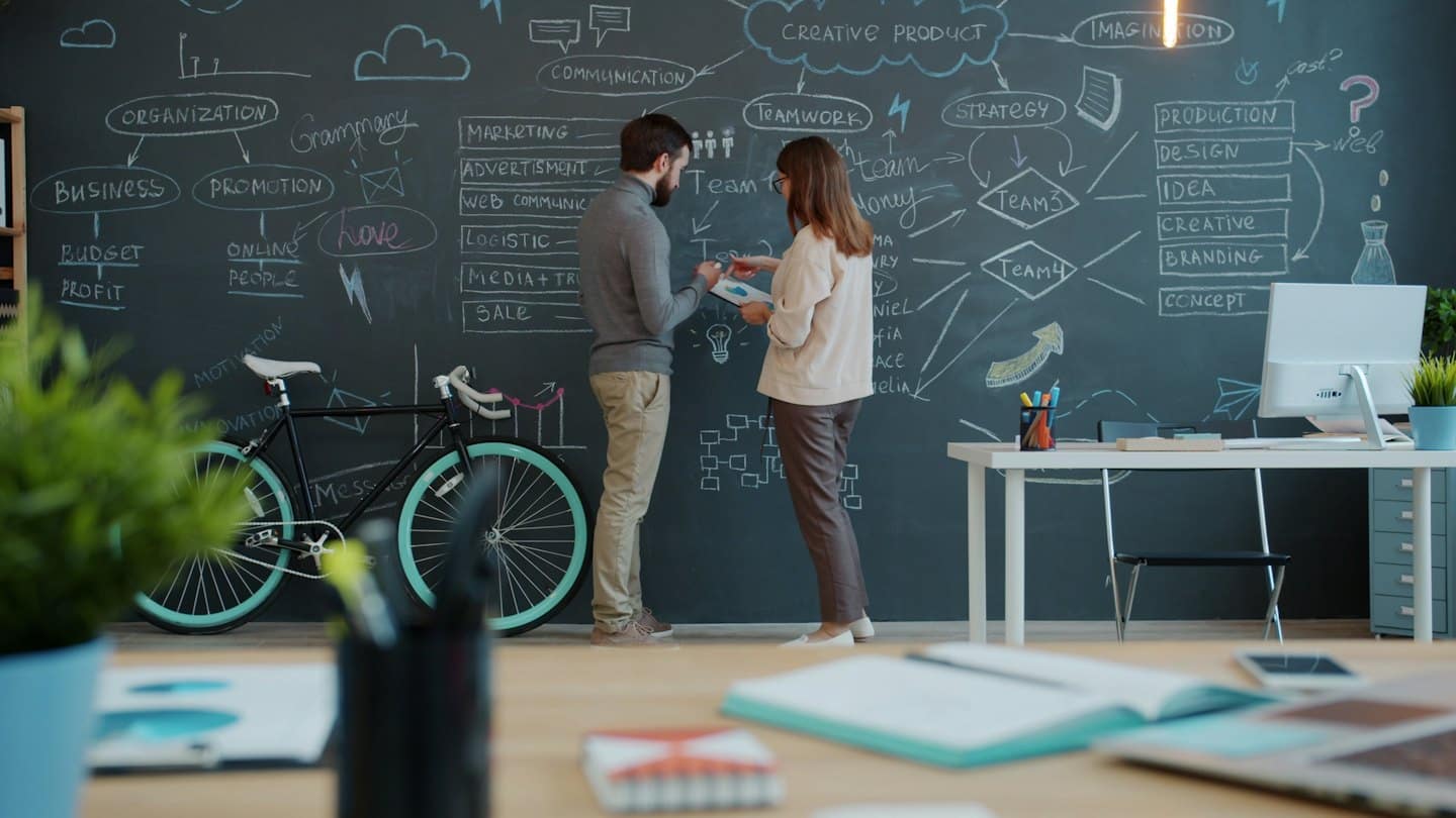 Two professionals discuss a strategy on a chalkboard in an office.